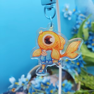 Puede incluir: Un amuleto de pez de dibujos animados dorado y naranja con un vestido azul y blanco. El amuleto está unido a una cuerda azul y blanca.
