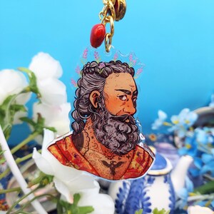 May include: A clear acrylic keyring depicting a detailed illustration of a bearded man with tattoos and a floral crown. The keyring is attached to a gold-coloured chain with a red bead charm. The background is a light blue.