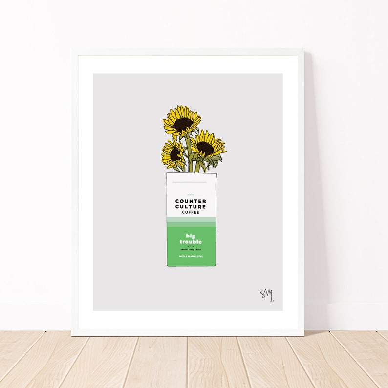 May include: A white framed print of a digital illustration of a bag of Counter Culture Coffee's "big trouble" blend with three sunflowers coming out of the top of the bag.