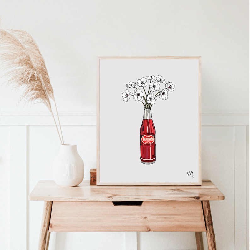 Może przedstawiać: Biały wydruk w ramie przedstawiający czerwoną szklaną butelkę z białymi kwiatami w środku. Butelka ma czerwoną etykietę z napisem "Dreamwhip". Wydruk jest podpisany "SM" w prawym dolnym rogu.