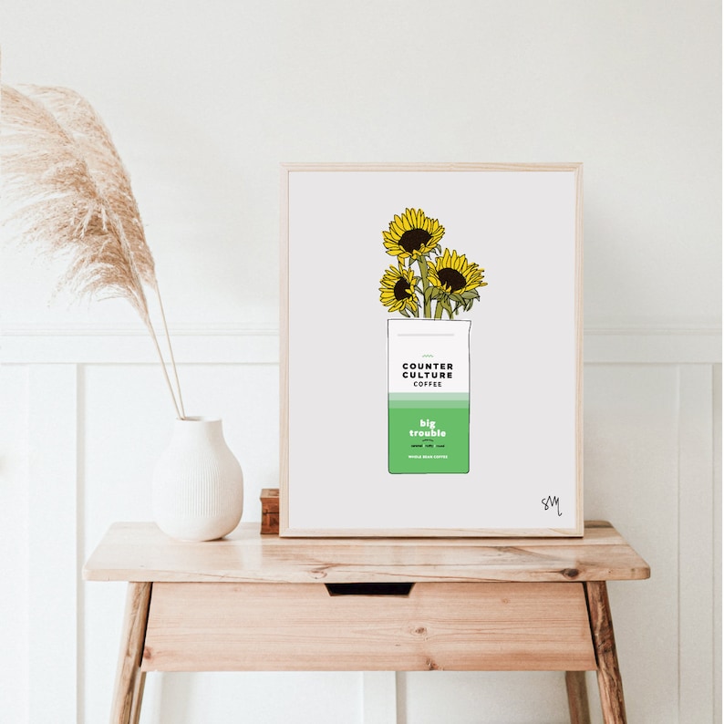 May include: A white framed print with a drawing of a bag of Counter Culture Coffee with sunflowers on top. The bag is green and has the text "Counter Culture Coffee" and "Big Trouble" on it.