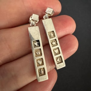 May include: A pair of silver drop earrings featuring a long rectangular bar design. Each earring displays four square indentations containing embossed symbols of a bird, a crown, a heart, and a trident-like shape, finished with a square stud top.