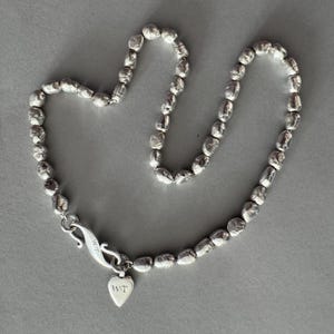 May include: A silver-toned necklace composed of numerous small, irregularly shaped, textured beads. The necklace features a curved metal hook clasp and a small, heart-shaped pendant dangling from the clasp, both engraved with the letters W.T.
