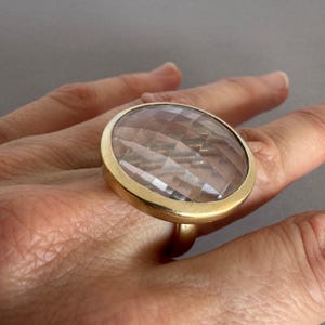 May include: A gold-toned ring featuring a large, circular, faceted clear stone set in a smooth metal bezel. The stone has a geometric, multi-faceted surface that reflects light, and the ring band is simple and solid, encircling the finger.
