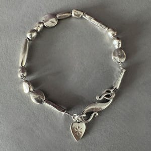 Könnte beinhalten: Ein silberfarbenes Armband mit einer Reihe unregelmäßiger, organisch geformter Perlen und länglicher zylindrischer Glieder. Das Schmuckstück verfügt über einen geschwungenen S-Haken-Verschluss und einen kleinen herzförmigen Anhänger mit einem Stern.