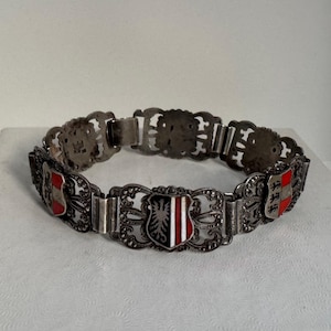 May include: Silver bracelet with decorative filigree links. Each link features a shield design with red, black, and white enamel. A vintage accessory.