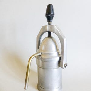 Può includere: Una macchina per caffè espresso manuale vintage. Il dispositivo è realizzato in metallo color argento con un manico nero e un beccuccio curvo in ottone. Ha una base cilindrica e una parte superiore a cupola, con una leva per premere.