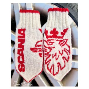 Könnte beinhalten: Ein Paar cremeweiße Strickhandschuhe mit roten Akzenten. Auf einem Handschuh steht "SCANIA" in Rot, auf dem anderen ein rotes Emblem. Die Bündchen und Kanten sind rot eingefasst.