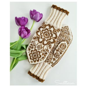 May include: A pair of cream and brown knit mittens featuring a detailed floral pattern and ribbed cuffs with brown trim, accompanied by two purple tulips with green stems and leaves. The text Småtterier is visible in the bottom right corner.