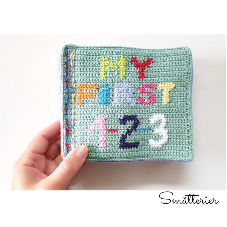 May include: A green crocheted square with the words "My First 1-2-3" in colorful yarn. The square is bordered with a multi-colored crocheted trim.