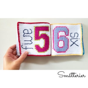 May include: A crocheted book with colorful numbers and the words "five" and "six" on the pages. The book is made of white yarn with red, yellow, and blue borders.