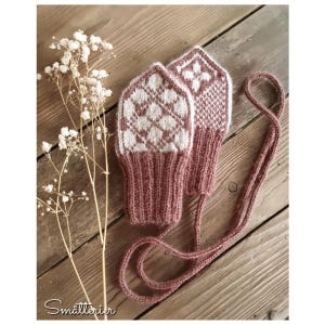 May include: A pair of dusty rose and white knit mittens featuring a geometric diamond pattern on the hand and a ribbed cuff. The mittens are connected by a long, matching dusty rose knitted cord. A sprig of white baby's breath flowers is nearby.