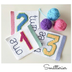 May include: Four crocheted squares with the numbers one, two, and three written on them. The squares are white with colored borders and the numbers are in different colors. There are three balls of yarn in the background.