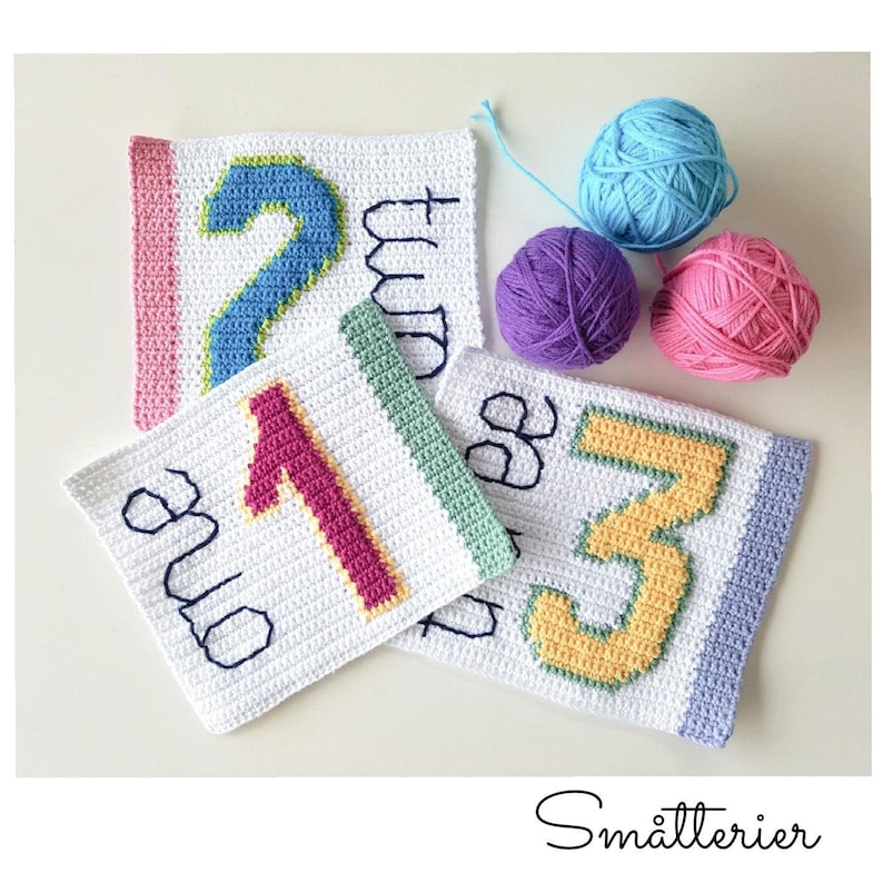 May include: Four crocheted squares with the numbers one, two, and three written on them. The squares are white with colored borders and the numbers are in different colors. There are three balls of yarn in the background.