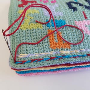 May include: Close-up of a crocheted blanket with a colorful design. The blanket is being stitched together with a red thread and a needle. The blanket has a multi-colored border.
