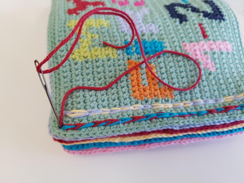 May include: Close-up of a crocheted blanket with a colorful design. The blanket is being stitched together with a red thread and a needle. The blanket has a multi-colored border.