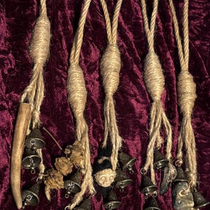 Pode incluir: Cinco borlas de corda castanha com pequenos sinos pendurados na parte inferior. Cada borla tem um pequeno objeto redondo castanho na parte superior. Uma borla tem um pequeno objeto castanho com forma humana na parte superior. As borlas est&atilde;o penduradas num fundo de veludo vermelho escuro.