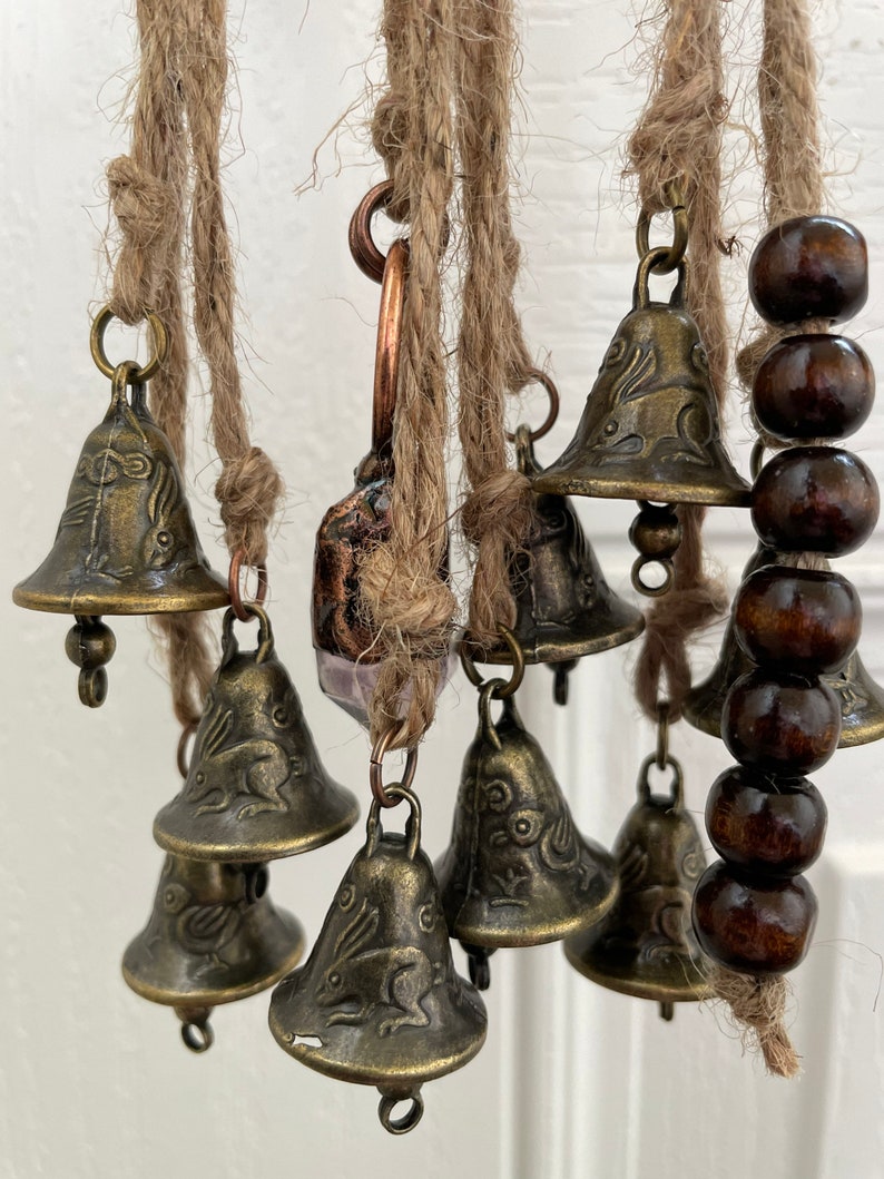 Pode incluir: Um m&oacute;bile de vento com v&aacute;rias pequenas sinos, cada um com um desenho de coelho, pendurados em um barbante marrom. Os sinos s&atilde;o de metal e t&ecirc;m um acabamento bronze. Tamb&eacute;m h&aacute; contas de madeira marrom penduradas no barbante.