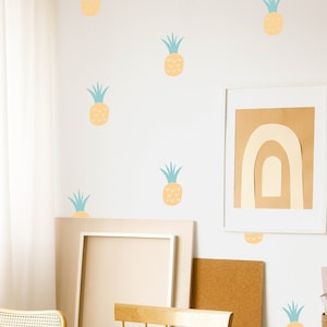 May include: A white wall with a light brown wooden table and two chairs. The wall is decorated with yellow pineapple stickers. There is a framed picture on the wall with a rainbow design.