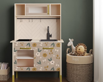Può includere: Una cucina giocattolo in legno con un design a fantasia floreale. La cucina ha un lavandino, un piano cottura e armadietti. La cucina è su un pavimento in legno e c'è un cesto di vimini con la parola "Giocattoli" su di esso.