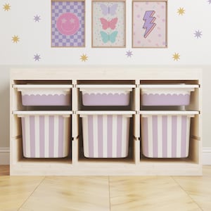 May include: A wooden storage unit with six compartments. The top three compartments hold lavender-colored bins with scalloped edges. The bottom three compartments hold bins with lavender and white vertical stripes. Above the unit are three framed prints with whimsical designs.