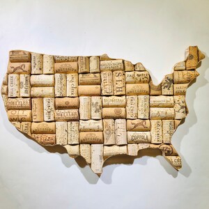 May include: A map of the United States crafted from numerous wine corks. The corks are various shades of brown and beige, with printed text and designs. The map is set against a white background, showcasing a rustic, handcrafted aesthetic.