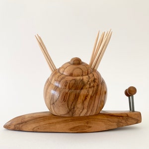 Puede incluir: Un porta palillos de dientes de madera con forma de caracol con un acabado de madera marrón. El caracol tiene un cuerpo redondo y una cola larga y fina. El porta palillos de dientes está lleno de palillos de dientes de madera.