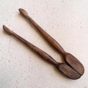 Puede incluir: Dos pinzas de cocina de madera con acabado marrón oscuro. Las pinzas tienen mangos largos y delgados y extremos redondeados. Un mango tiene crestas talladas para el agarre. Las pinzas están diseñadas para recoger y servir alimentos.