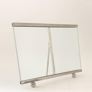 Puede incluir: Un marco de fotos rectangular de metal plateado y cristal. El marco tiene un diseño sencillo con una parte delantera de cristal transparente y una parte trasera de metal. Se mantiene en posición vertical con un pequeño soporte metálico y dos pies esféricos.