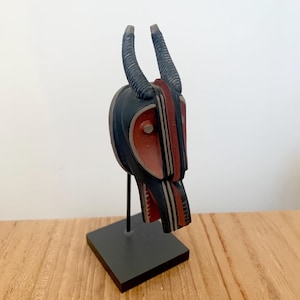 Puede incluir: Escultura de madera tallada de una cabeza de animal con cuernos curvos. La escultura presenta una combinación de colores negro, rojo y crema. Está montada sobre un soporte de metal negro con una base cuadrada. La obra de arte se exhibe sobre una superficie de madera.