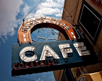 May include: A vintage neon sign for a cafe with the word "CAFE" in white neon letters and the words "COCKTAILS" in red neon letters. The sign is mounted on a building with a blue sky and white clouds in the background.