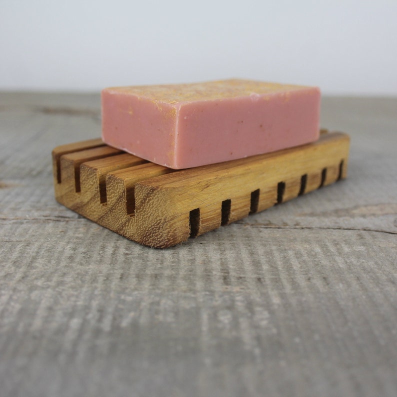 May include: A wooden soap dish with a pink bar of soap on top. The soap dish has a slatted design to allow water to drain.
