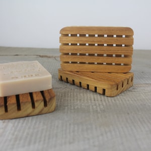 May include: Two wooden soap dishes made of teak wood. The top dish has a slatted design and the bottom dish has a solid design. A bar of soap is sitting on the top dish.