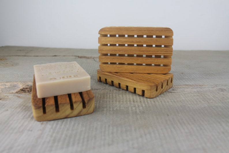 May include: Two wooden soap dishes made of teak wood. The top dish has a slatted design and the bottom dish has a solid design. A bar of soap is sitting on the top dish.