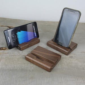Könnte beinhalten: Drei Holz-Handyständer mit dunkelbrauner Oberfläche. Zwei Ständer haben Smartphones, einer mit einem Samsung Galaxy Note7 Bildschirm, der die Zeit 12:45 anzeigt. Der dritte Ständer ist leer.