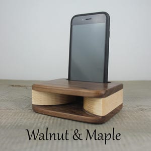 Puede incluir: Un soporte de tel&eacute;fono de madera con acabado en nogal y arce. El soporte tiene un amplificador incorporado para mejorar el sonido. Un tel&eacute;fono inteligente negro est&aacute; colocado en el soporte.