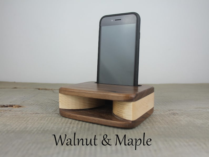 Puede incluir: Un soporte de tel&eacute;fono de madera con acabado en nogal y arce. El soporte tiene un amplificador incorporado para mejorar el sonido. Un tel&eacute;fono inteligente negro est&aacute; colocado en el soporte.