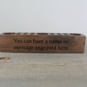 May include: A wooden pencil holder with multiple holes for pencils and a large space for other items. The holder is made of dark wood and has the text "You can have a name or message engraved here" on the front.