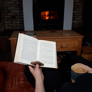 May include: A person is sitting in front of a fireplace reading a book with a wooden page holder. They are holding a cup of coffee in their other hand. The fireplace is lit and there is a wooden table in front of it.
