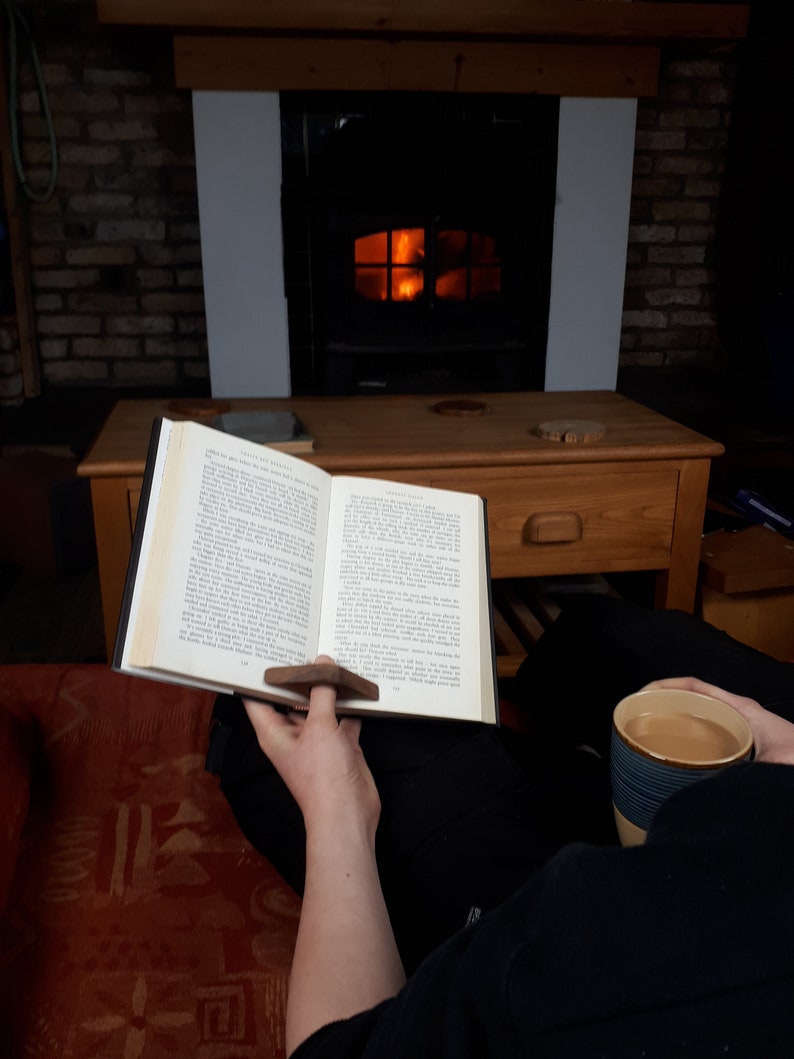 May include: A person is sitting in front of a fireplace reading a book with a wooden page holder. They are holding a cup of coffee in their other hand. The fireplace is lit and there is a wooden table in front of it.