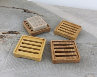 Puede incluir: Cuatro jaboneras cuadradas de madera con diseño de listones. Una jabonera contiene una pastilla de jabón con texto. Los tonos de madera varían de marrón claro a oscuro. La jabonera está diseñada para permitir el drenaje del agua.