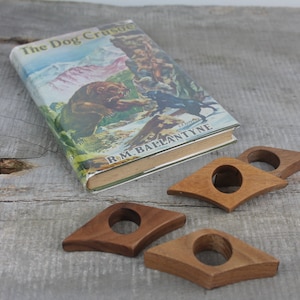 May include: Four wooden book page holders in the shape of a teardrop. The holders are brown and have a hole in the center. The holders are on a wooden surface with a book titled "The Dog Crusoe" by R.M. Ballantyne.
