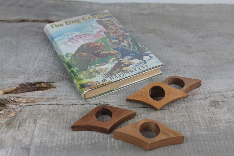 May include: Four wooden book page holders in the shape of a teardrop. The holders are brown and have a hole in the center. The holders are on a wooden surface with a book titled "The Dog Crusoe" by R.M. Ballantyne.