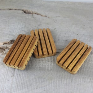 May include: Three wooden soap dishes with grooved bottoms. The dishes are made of light brown wood and are arranged in a row on a gray wooden surface.