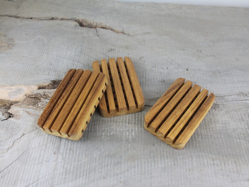 May include: Three wooden soap dishes with grooved bottoms. The dishes are made of light brown wood and are arranged in a row on a gray wooden surface.
