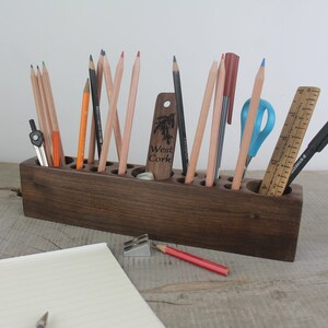 May include: A wooden pencil holder with 12 compartments, filled with pencils, pens, a ruler, a compass, and scissors. The holder is stained a dark brown color and has the words "West Cork" engraved on the front.