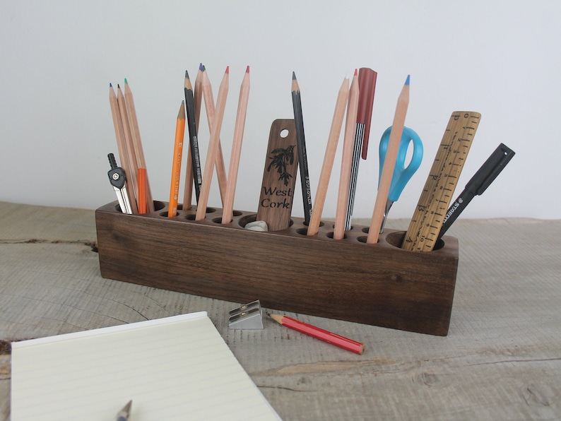 May include: A wooden pencil holder with 12 compartments, filled with pencils, pens, a ruler, a compass, and scissors. The holder is stained a dark brown color and has the words "West Cork" engraved on the front.