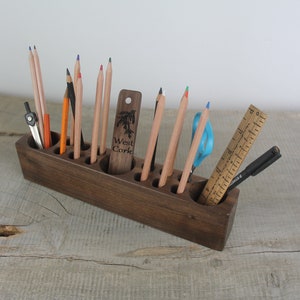 May include: A dark brown wooden pencil holder with 10 slots for pencils, pens, and other writing tools. The holder is filled with colored pencils, a compass, a ruler, a pen, and a pair of scissors. The holder has the text "West Cork" engraved on the side.