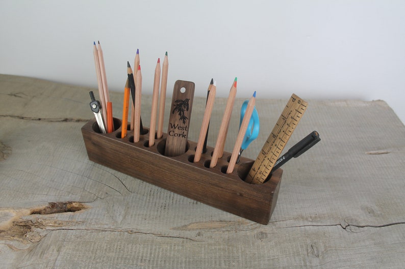 May include: A dark brown wooden pencil holder with 10 slots for pencils, pens, and other writing tools. The holder is filled with colored pencils, a compass, a ruler, a pen, and a pair of scissors. The holder has the text "West Cork" engraved on the side.