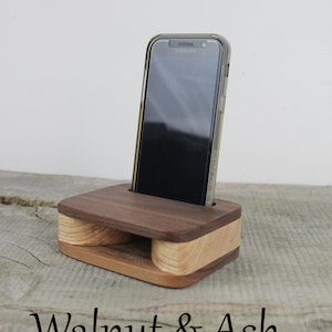 Puede incluir: Soporte para tel&eacute;fono de madera de nogal y fresno con un tel&eacute;fono inteligente negro en su interior. El soporte tiene un recorte para amplificar el sonido.